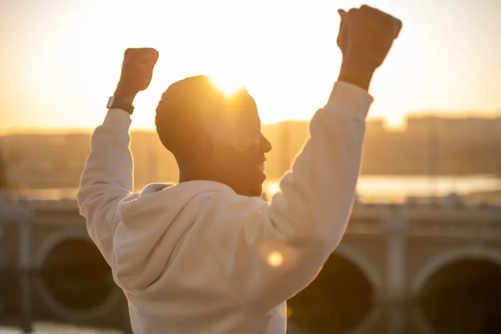 Man lifting both arms toward a warm sunset above the city